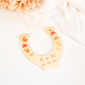 May include: A cream-coloured ceramic horseshoe ornament featuring hand-painted orange and red maple leaves along the sides. The centre is personalised with the letters R & M and the date 01 15 24 in a reddish-brown font, suspended by a thin ribbon.