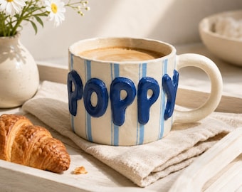 Può includere: Una tazza in ceramica con base color crema, strisce verticali azzurre e la parola POPPY scritta in lettere arrotondate, in rilievo e di colore blu scuro. La tazza ha un manico curvo e contiene un liquido marrone su un vassoio chiaro.