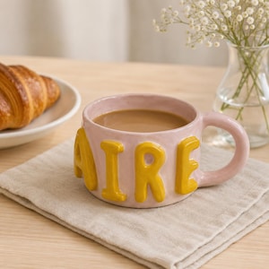 May include: A light pink ceramic mug with a rounded handle and a textured surface. The mug features large, raised, mustard yellow letters spelling out the word AIRE around the exterior. The mug is filled with a light brown liquid beverage.