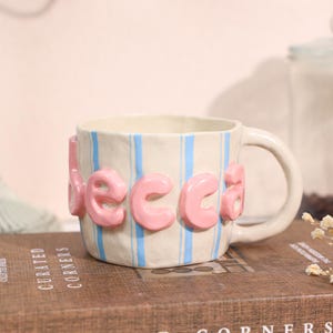 May include: A ceramic mug with a cream-coloured base featuring vertical light blue stripes. The mug has a thick, rounded handle and large, raised, bubble-style pink letters spelling out the name Becca wrapped around the exterior surface.