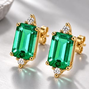 May include: A pair of gold stud earrings featuring a large rectangular emerald-cut green gemstone in the center, accented by two small round white diamonds at the top and bottom points of each setting, secured with gold post backings.