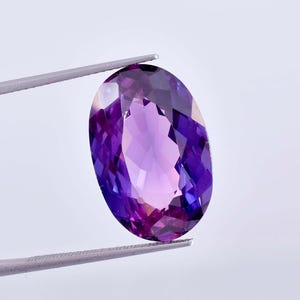 May include: An oval-cut, faceted gemstone with a vibrant purple and violet colour gradient. The stone is held by a pair of metal tweezers, showcasing its translucent quality and geometric facets that reflect light across its polished surface.