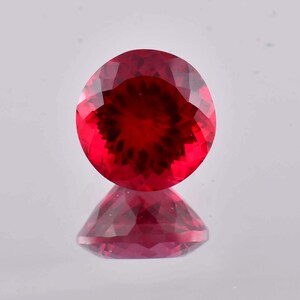 Puede incluir: Una piedra preciosa redonda y facetada de un color rojo intenso y vibrante. La piedra presenta un corte preciso con numerosas facetas triangulares que crean un patrón simétrico en forma de estrella de reflexión de luz en su superficie.