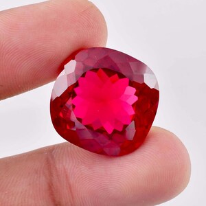 Puede incluir: Una gema roja de talla cojín con una superficie altamente facetada que crea un patrón de reflexión de luz en forma de estrella. La piedra muestra un tono carmesí profundo y saturado, sostenida para exhibir su claridad y corte.