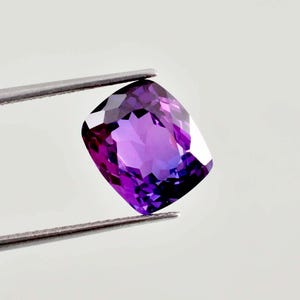 May include: A cushion-cut gemstone with a deep purple hue and multifaceted surface held by silver-toned metal tweezers. The stone displays varying shades of violet and dark purple, reflecting light across its polished, geometric, and symmetrical facets.