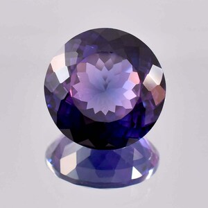 May include: A round, faceted gemstone displaying a deep purple and violet colour gradient. The stone features a complex, symmetrical cut with numerous triangular and diamond-shaped facets that create a star-like pattern in the centre.