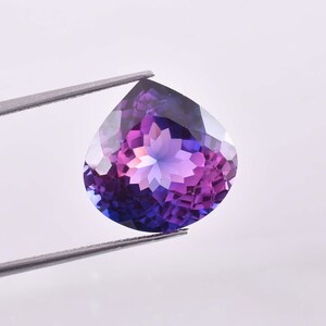 May include: A pear-shaped faceted gemstone displaying a vibrant gradient of deep purple and blue hues. The stone features a complex geometric cut with numerous triangular facets that reflect light, held by a pair of thin metal tweezers.