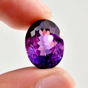 May include: An oval-cut amethyst gemstone featuring a deep purple hue with lighter violet facets. The stone displays a brilliant, multi-faceted surface that reflects light, showcasing a clear, transparent quality and a symmetrical, polished appearance.