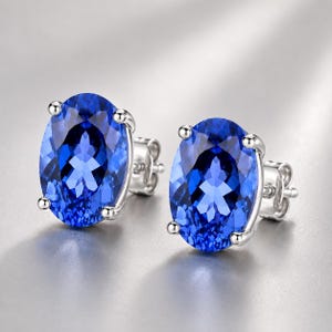 May include: A pair of stud earrings featuring large, oval-cut, vibrant blue gemstones set in polished silver-toned metal. Each stone is secured by four prongs, showcasing a faceted surface that reflects light, with visible metal post backings.