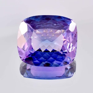 May include: A cushion-cut gemstone displaying a vibrant gradient of deep blue and soft purple hues. The stone features a complex faceted surface that creates sharp geometric reflections and a high level of clarity throughout its transparent body.