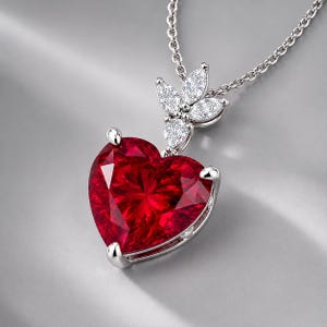 Puede incluir: Un colgante de piedra preciosa roja en forma de corazón engastado en plata, con un ajuste de tres puntas y un pasador adornado con tres piedras claras de corte marquesa. El colgante cuelga de una delicada cadena de plata con acabado metálico.