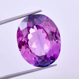May include: An oval-cut gemstone with a vibrant purple hue and multifaceted surface that reflects light. The stone is held by metal tweezers, showcasing its transparent clarity and deep violet colour tones throughout the polished, symmetrical facets.