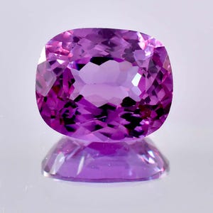 Puede incluir: Una piedra preciosa de corte cojín con un tono púrpura vibrante y una superficie facetada que refleja la luz. La piedra muestra una calidad clara y transparente con patrones geométricos internos creados por los cortes precisos realizados.
