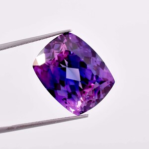 May include: A cushion-cut gemstone with a vibrant purple and violet colour gradient. The stone features a faceted surface that reflects light, creating a geometric pattern of deep and light purple hues throughout the transparent crystalline structure.
