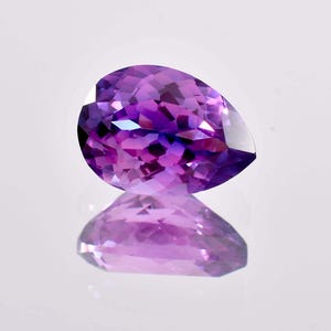 May include: A pear-shaped amethyst gemstone featuring a faceted cut that creates a brilliant display of deep purple and violet hues. The stone exhibits a translucent quality with sharp geometric facets that reflect light across its surface.