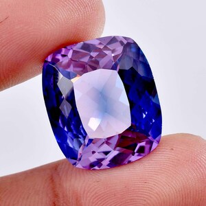 May include: A cushion-cut gemstone displaying a vibrant colour gradient that transitions from deep royal blue at the edges to a lighter, translucent lavender purple in the centre, featuring a faceted surface that reflects light across its geometric planes.