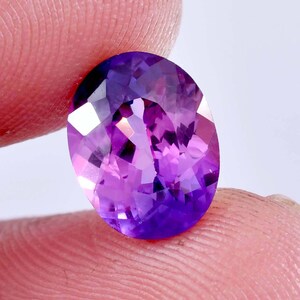 May include: An oval-cut gemstone with a vibrant purple hue and multifaceted surface that reflects light. The stone displays deep violet tones and lighter lavender highlights throughout its transparent structure, showcasing a polished finish.