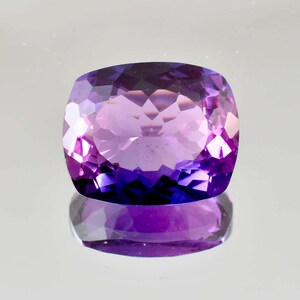 May include: A cushion-cut amethyst gemstone featuring a vibrant purple hue with deep violet saturation. The stone displays a faceted surface that creates intricate geometric light reflections and a clear mirror-like reflection on the surface below.