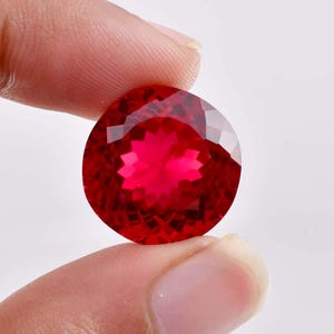 Puede incluir: Una piedra preciosa redonda y facetada de un color rojo intenso y vibrante. La piedra presenta un corte preciso de múltiples facetas que crea un patrón de reflejos de luz en forma de estrella en su superficie, mostrando gran claridad.