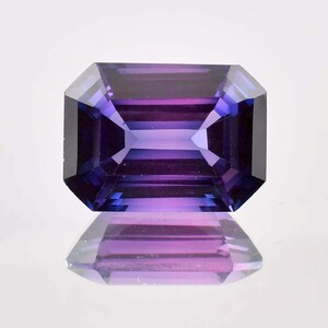 May include: A loose emerald-cut gemstone featuring a deep purple and violet colour gradient. The stone has a faceted surface with sharp, geometric edges and a clear, transparent quality that allows light to pass through, creating a distinct reflection.