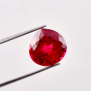 Op de afbeelding: Een levendige, dieprode edelsteen met een gefacetteerde, peervormige slijpvorm. De steen vertoont een hoge helderheid en schittering en reflecteert het licht door de vele geometrische vlakken. De steen wordt vastgehouden door een zilverkleurige pincet.