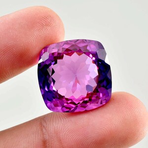 May include: A cushion-cut gemstone featuring a vibrant gradient of deep purple and bright magenta hues. The stone has a faceted surface that creates a complex geometric pattern of light and shadow, reflecting a brilliant, multi-toned appearance.