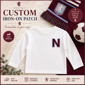 Puede incluir: Una camiseta blanca de manga larga con un parche termoadhesivo personalizado de la letra N en color granate con un borde azul claro. La prenda se muestra junto a una bufanda del Aston Villa y un balón de fútbol.