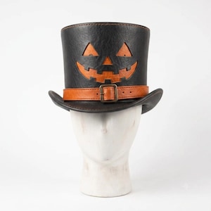 Puede incluir: Un sombrero de copa negro con superficie texturizada y un diseño recortado de cara de calabaza de Halloween. El sombrero incluye una banda naranja en la base con una hebilla metálica, revelando una capa naranja bajo el material exterior.