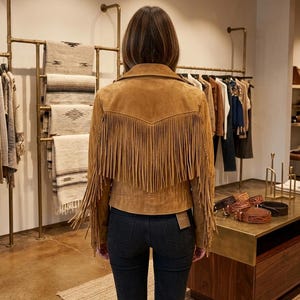 Pode incluir: Uma jaqueta de camur&ccedil;a bege com franjas longas no jugo traseiro e ao longo das mangas. A jaqueta possui gola e &eacute; usada com cal&ccedil;as jeans cinza-escuras. Uma pequena etiqueta retangular est&aacute; fixada na bainha inferior da pe&ccedil;a.