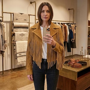 Pode incluir: Uma jaqueta de camur&ccedil;a bege com franjas longas nas mangas e pain&eacute;is frontais, combinada com uma camiseta branca lisa e cal&ccedil;a jeans cinza escuro. A jaqueta tem uma gola larga e um corte curto com fechamento frontal por bot&otilde;es.