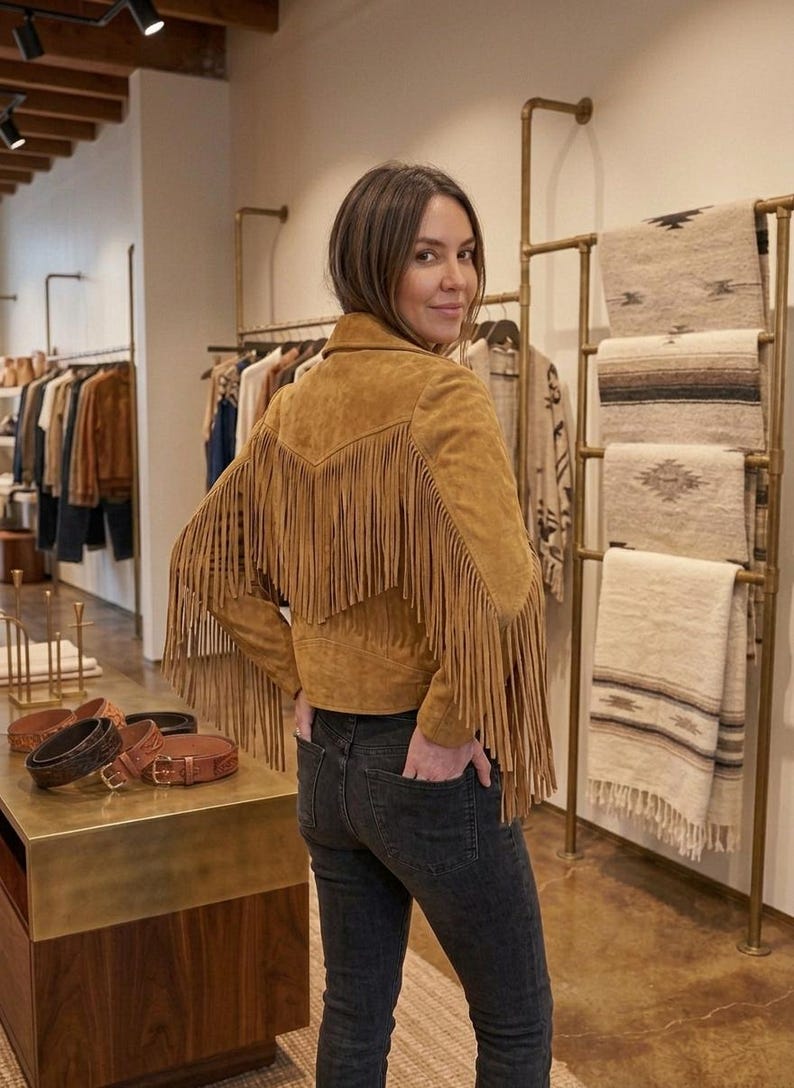 Pode incluir: Uma jaqueta de camur&ccedil;a bege com longas franjas nas costas e nas mangas, combinada com cal&ccedil;as jeans cinza-escuras. A jaqueta tem gola e silhueta ajustada, destacando o material texturizado e um design cl&aacute;ssico inspirado no estilo western.