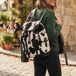 May include: A backpack featuring a black and white cow print pattern with black leather trim, adjustable straps, and two side pockets with buckle closures. The bag has a top flap with a drawstring closure and vertical leather straps with buckles.
