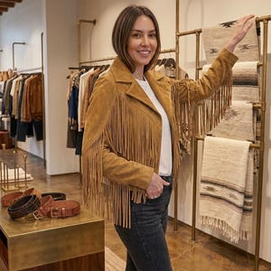 Pode incluir: Uma jaqueta de camur&ccedil;a bege com longas franjas ao longo das mangas e nos pain&eacute;is frontais. A jaqueta tem uma gola cl&aacute;ssica, fecho frontal com z&iacute;per e &eacute; usada sobre uma camiseta branca lisa combinada com cal&ccedil;as jeans cinza escuro.