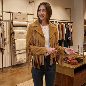 Pode incluir: Uma jaqueta de camur&ccedil;a bege com franjas longas nas mangas e pain&eacute;is frontais, combinada com uma camiseta branca lisa e cal&ccedil;a jeans cinza escuro. A jaqueta tem uma gola larga e um corte curto com um acabamento texturizado.