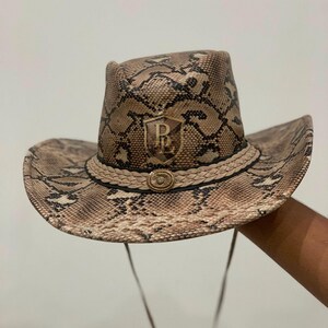Puede incluir: Un sombrero de vaquero con un patrón de piel de serpiente en tonos beige y negro. El sombrero incluye una banda trenzada de color beige con un emblema metálico central y una placa en forma de escudo con las letras PL en acabado dorado.