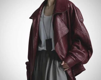Pode incluir: Um casaco de couro bordeaux oversize com gola larga, mangas compridas e presilhas abotoadas, usado sobre uma t-shirt branca lisa e calças cinzentas. Um colar de cordão preto fino com um pequeno pendente retangular ao pescoço.