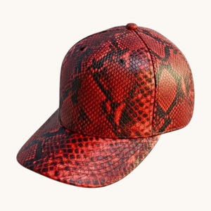 Peut inclure: Une casquette de baseball arborant un motif peau de serpent rouge et noir vif sur toute sa surface. La casquette présente une calotte structurée, une visière incurvée et un bouton au sommet. L'imprimé texturé imite l'aspect des écailles.