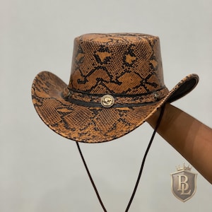 Puede incluir: Un sombrero de vaquero con un patrón de piel de serpiente en marrón y negro. El sombrero incluye una banda negra con textura trenzada y un emblema central circular de metal dorado. Una fina correa de barbilla marrón cuelga de los lados.