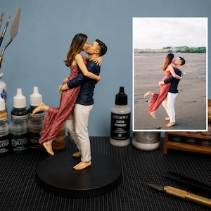 May include: A custom 3D figurine depicting a couple in an embrace, with one person wearing a long red patterned dress and the other in a dark blue long-sleeved shirt and white trousers, standing on a circular black base, modeled after a photograph.