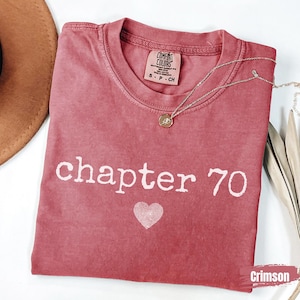 May include: A crimson short-sleeve t-shirt with a crew neckline featuring the white text chapter 70 and a white fingerprint heart graphic. The inner neck label reads Comfort Colors, 100% cotton, made in Honduras, size S P CH.