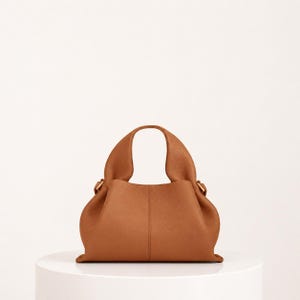 May include: A brown leather handbag with a rounded shape and a single handle. The bag has a structured base and a soft, gathered body. Gold-colored hardware accents the handle attachments. The bag is displayed on a white surface.