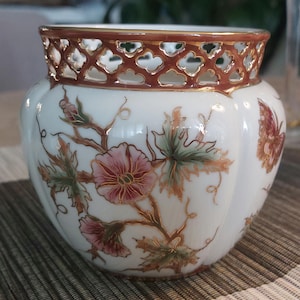 Può includere: Un vaso in ceramica bianca con un motivo floreale. Il vaso presenta fiori rosa, foglie verdi e un bordo a griglia marrone. C'è anche una farfalla dipinta sul lato. È un oggetto decorativo.