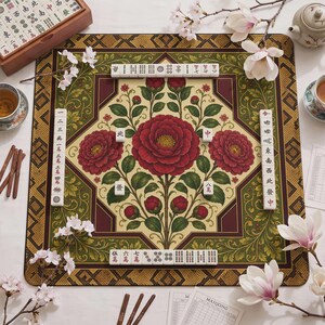 May include: A square mahjong mat featuring a central cream-colored panel with large red camellia flowers and green foliage, surrounded by a dark green and gold geometric border. White mahjong tiles with black and red characters are arranged on the mat.