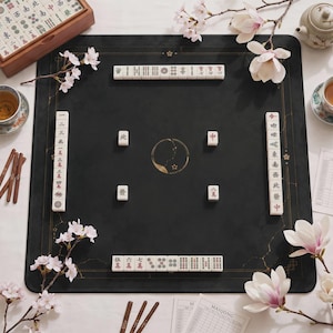 May include: A square black mahjong mat featuring gold-colored line borders and a central circular emblem. The mat is arranged with white mahjong tiles displaying black and red characters, accompanied by cherry blossom branches and a score sheet.