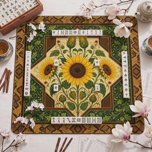 May include: A square mahjong mat featuring a central illustration of yellow sunflowers with green leaves on a cream background, framed by a dark green and brown border with geometric patterns. White mahjong tiles are arranged along the four sides.