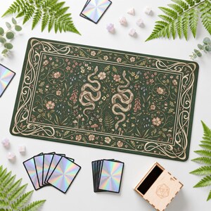 Könnte beinhalten: Eine rechteckige Spielmatte mit dunkelgrünem Hintergrund, die zwei ineinander verschlungene Schlangen inmitten botanischer Illustrationen von Blumen, Blättern und Pilzen zeigt. Das Design ist von einem cremefarbenen Rand mit Blumenmotiven umgeben.