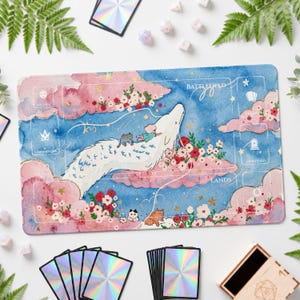 Pode incluir: Um tapete de jogo retangular com uma ilustração em aguarela de um dragão branco a voar através de nuvens cor-de-rosa e um céu azul. O design inclui detalhes florais, uma pequena figura e gatos, com zonas de jogo impressas como Battlefield, Lands e Graveyard.
