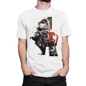 May include: A white short-sleeved t-shirt featuring a detailed black and white graphic illustration of a character with an eye patch and a mechanical hand, accented with vibrant red highlights on the hand and surrounding elements.