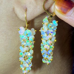 May include: A pair of drop earrings featuring clusters of small, round, iridescent white opals with flashes of green, blue, and pink. The stones are connected by thin gold-toned wire and suspended from gold-toned lever-back earring hooks.