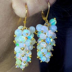 May include: A pair of drop earrings featuring clusters of small, round, iridescent white opal beads with gold-toned accents. Each earring is suspended from a gold-toned lever-back hook, creating a cascading, grape-like shape with colourful flashes.